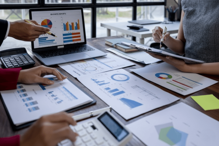 A close-up of professionals reviewing financial documents and colorful data charts on a wooden desk with a laptop and calculator.
