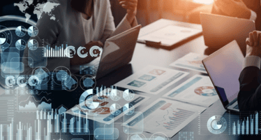 A high-angle shot captures a business meeting, where two individuals are seen collaborating while reviewing paperwork and engaging with their laptops.