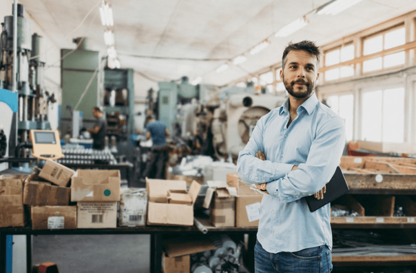 A man with a beard and a light blue shirt, holding a tablet, standing with arms crossed in an industrial factory with machines in the background.