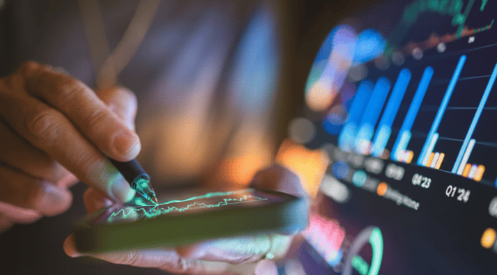 A person uses a stylus on a smartphone displaying a green line graph, with colorful data charts visible on a larger screen in the blurred background.