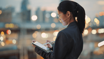 A young East Asian woman in a blazer stands outdoors at dusk, focused on using a stylus and tablet against a soft-focus city backdrop with glowing bokeh lights.