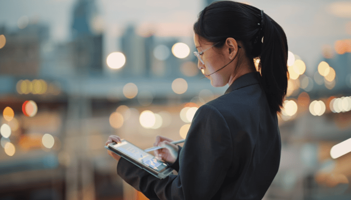 A young East Asian woman in a blazer stands outdoors at dusk, focused on using a stylus and tablet against a soft-focus city backdrop with glowing bokeh lights.