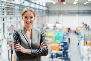 A smiling blonde woman in a grey blazer stands with crossed arms in a factory.