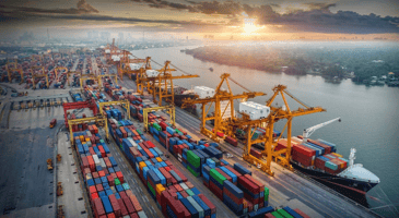 High-angle view of a bustling container terminal at sunset, featuring rows of colorful shipping containers, large yellow gantry cranes, and a cargo ship docked along a river.