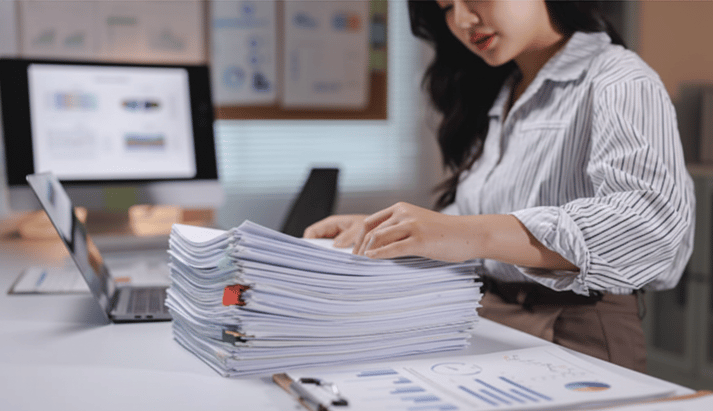 A young woman is organizing a tall stack of documents and paperwork on an office desk, next to a laptop and charts.