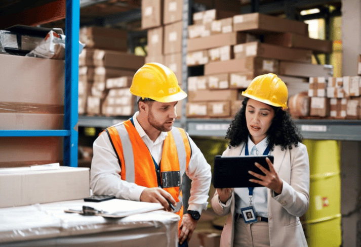 Two workers in hard hats examine a tablet in a warehouse.