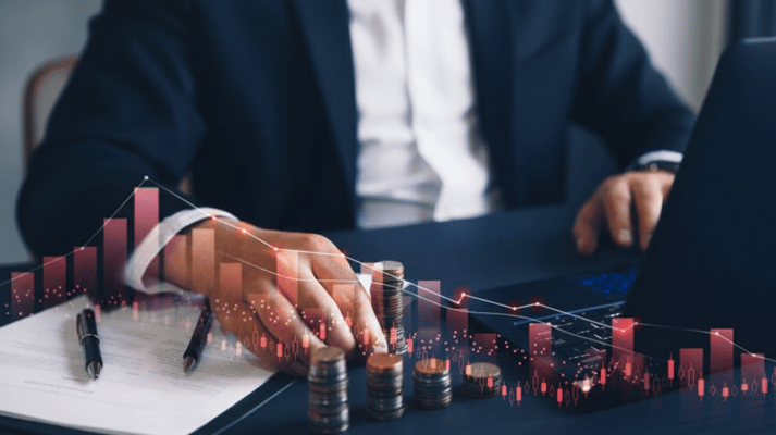 A person in a suit sits at a desk with stacks of coins and a laptop, overlaid with a translucent red financial graph.