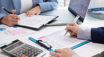 Two professionals reviewing financial spreadsheets and "Costing Reports" at a desk with a calculator and laptop.