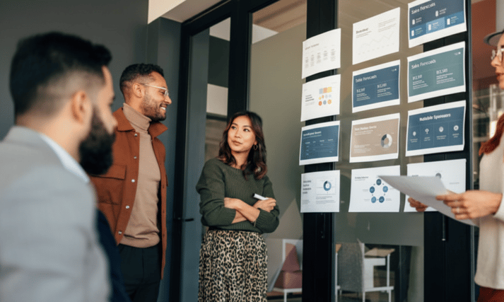 A diverse group of professionals engaging in a lively discussion in a modern office setting, with one person presenting information displayed on glass partitions behind them.