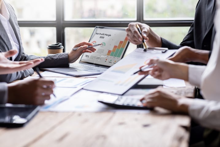 A group of business professionals collaborating around a table, analyzing financial reports with charts and graphs on a laptop and paper documents.