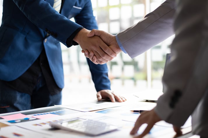 Two businessmen in suits shake hands over a table with documents.
