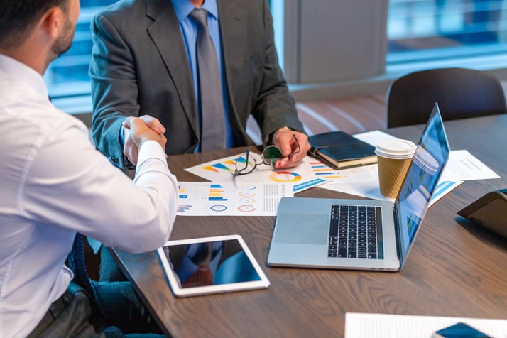 Two businessmen in formal attire shaking hands across a conference table, surrounded by charts, graphs, a laptop, tablet, notebook, and a coffee cup—symbolizing a successful business agreement.