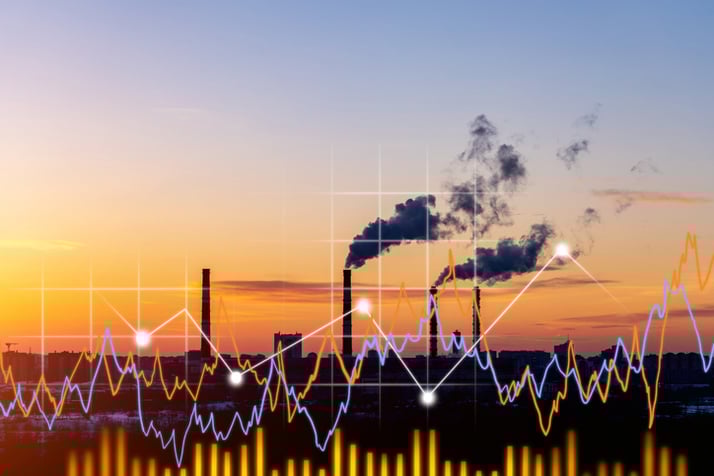 ndustrial factory at sunset with smoke rising from chimneys, overlaid with colorful financial graphs representing manufacturing performance data.