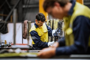 Two industrial workers wearing yellow and navy safety uniforms operate machinery in a manufacturing facility, with one worker in focus using a drill press and another blurred in the foreground