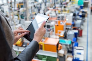 Person using a digital tablet to monitor operations inside a modern manufacturing facility with machinery and equipment in the background.