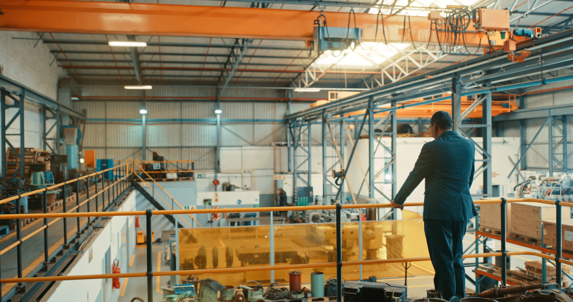 A businessman in a dark suit stands on an elevated walkway, overlooking machinery and equipment inside a large industrial manufacturing facility.