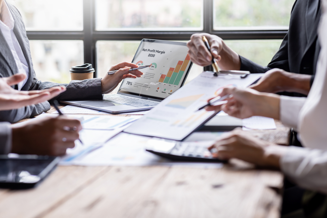 A group of business professionals collaborating around a table, analyzing financial reports with charts and graphs on a laptop and paper documents.