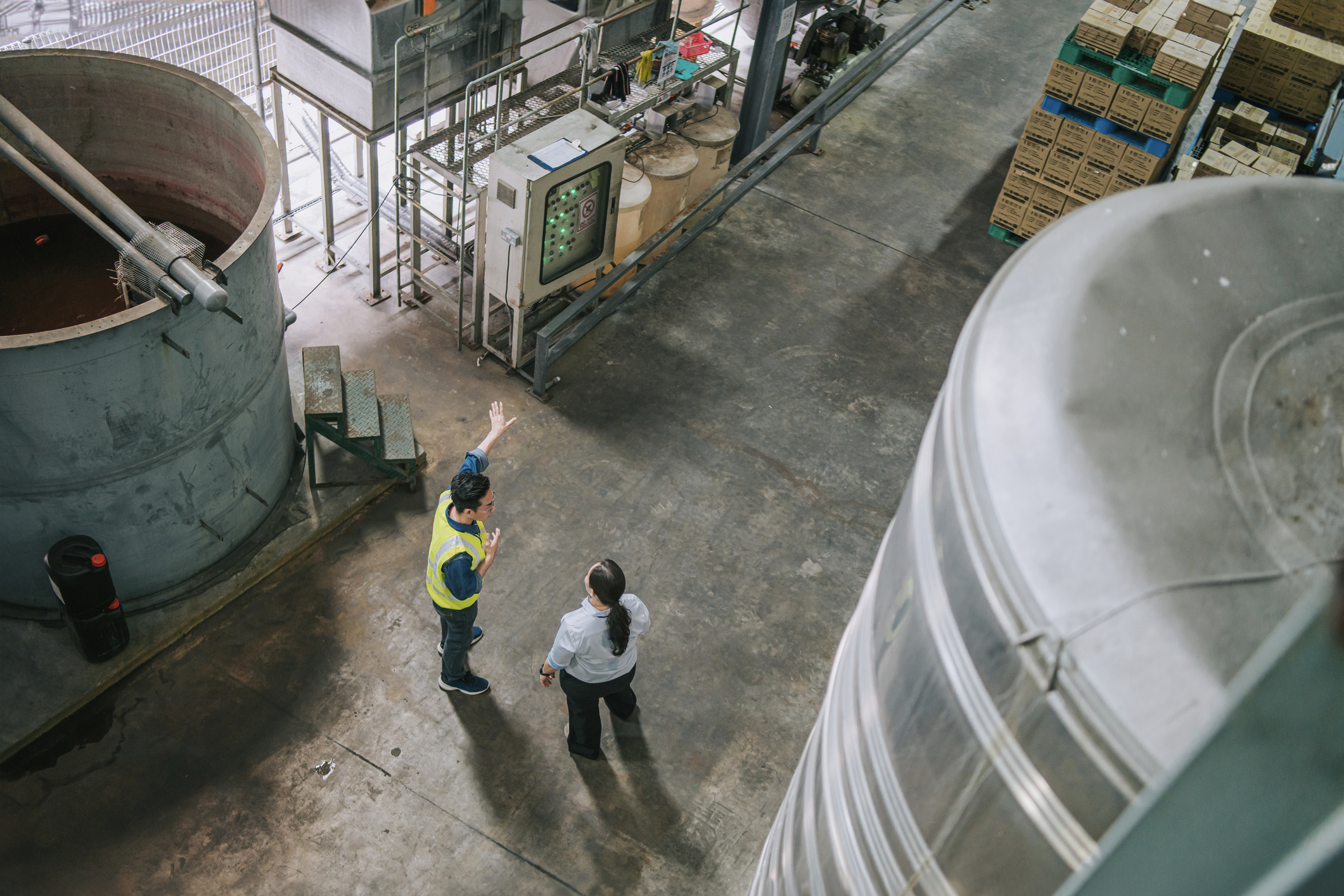 Overhead view of two people in safety attire discussing operations on a factory floor, surrounded by large industrial tanks, machinery, and neatly stacked boxes in a warehouse setting.