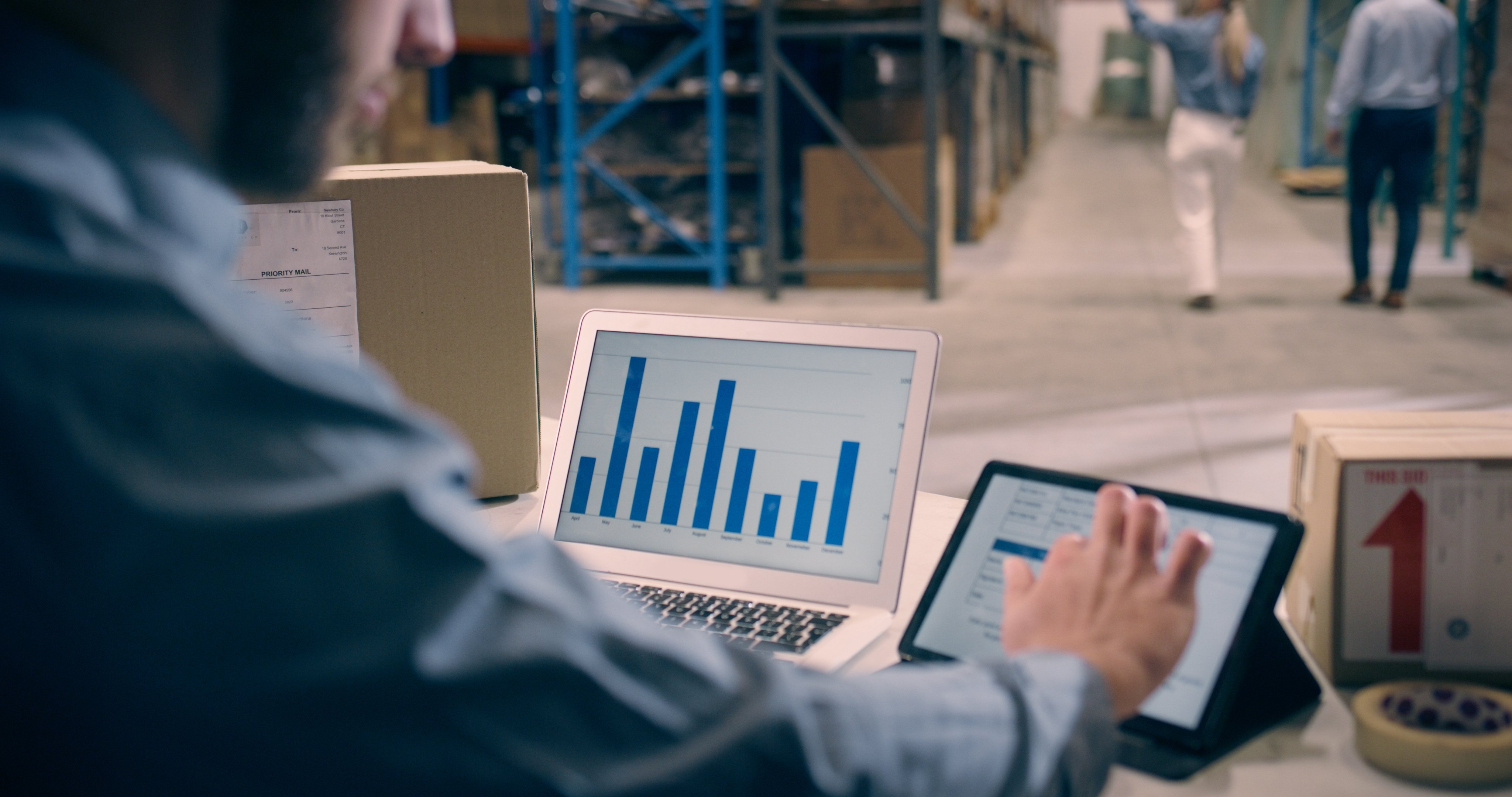 Person analyzing sales or inventory data on a laptop and tablet in a warehouse, with bar graphs displayed on screen and shipping boxes in the background.