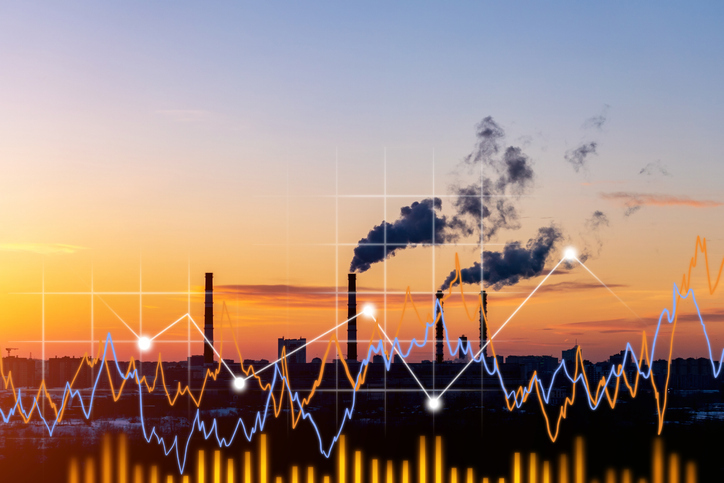 ndustrial factory at sunset with smoke rising from chimneys, overlaid with colorful financial graphs representing manufacturing performance data.