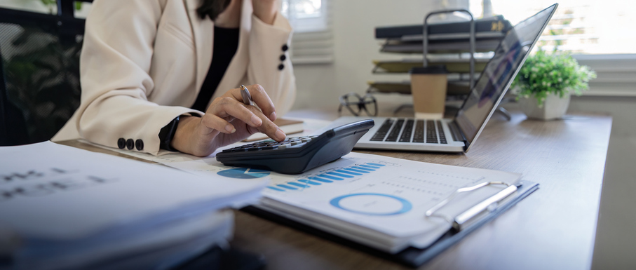 Business professional using a calculator and laptop to analyze financial data at a desk, with charts, documents, and a financial report in view.