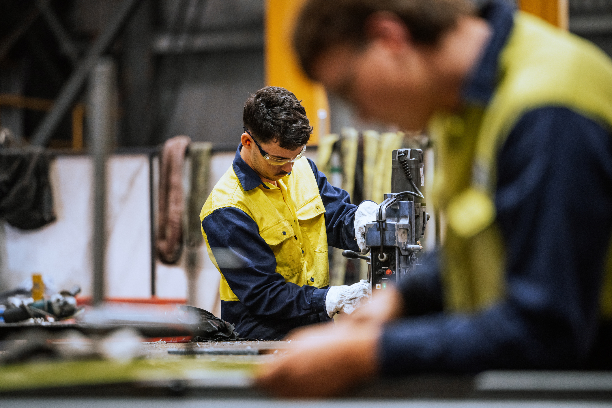 Two industrial workers wearing yellow and navy safety uniforms operate machinery in a manufacturing facility, with one worker in focus using a drill press and another blurred in the foreground