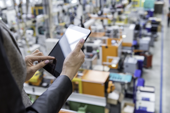 Person using a digital tablet to monitor operations inside a modern manufacturing facility with machinery and equipment in the background.