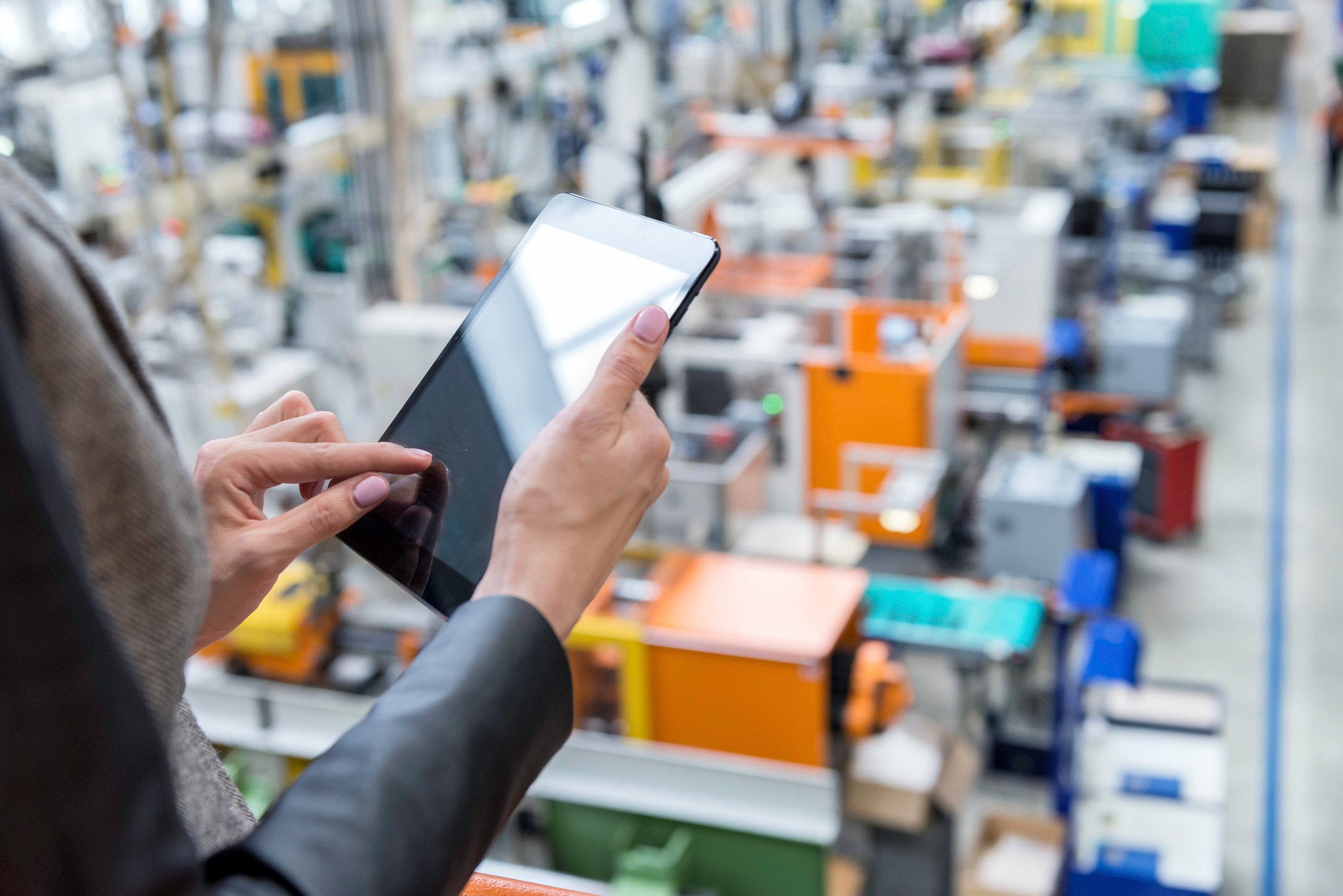 Person using a digital tablet to monitor operations inside a modern manufacturing facility with machinery and equipment in the background.